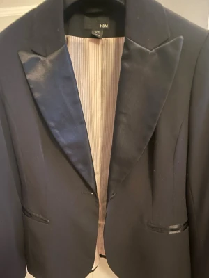 Svart kavaj med satindetaljer H&M - Stilren svart kavaj från H&M med blanka satinslag och matchande satindetaljer vid fickorna. Kavajen har en knapp framtill och är helfodrad med ljusrandigt tyg på insidan. Perfekt för en dressad look.