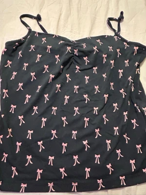 Svart linnetopp med rosa rosetter - Söt svart linnetopp med rosa rosett-mönster och knytbara axelband. Toppen har en lätt rynkning vid bysten som ger en fin form. Perfekt för dig som gillar gulliga detaljer och en avslappnad stil.