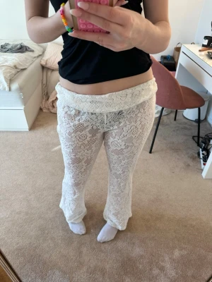 Vita spetsleggings med blommönster - Säljer ett par vita strandbyxor i genomskinlig spets med blommigt mönster och utsvängda ben. Byxorna har en bred, vikt linning och är perfekta att dra över bikinin på stranden. Materialet är lätt och luftigt, perfekt för sommardagar.