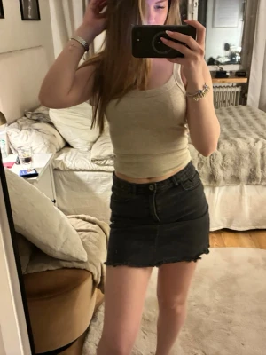 Svart jeanskjol med rå kant - Säljer en svart kort jeanskjol med rå, fransig nederkant och klassiska fickor fram och bak. Kjolen har en normal passform och stängs med knapp och dragkedja. Perfekt för dig som gillar en enkel och stilren look.