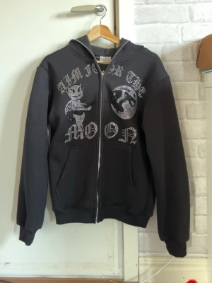 Aim for the moon - Svart hoodie med dragkedja och huva. Framsidan har ett stort tryck med texten 'I'M FROM THE MOON', en tecknad figur och en måne. Hoodien har två fickor fram och ribbade muddar. Perfekt för en cool och avslappnad stil.