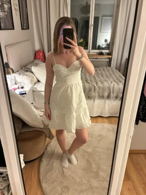 Vit kort klänning med volanger - Söt vit klänning med volanger och smala axelband. Klänningen har en v-ringad bustierliknande överdel och är kort i modellen. Perfekt för sommarens alla tillfällen. Materialet känns lätt och luftigt.