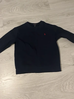 Mörkblå sweatshirt från Polo Ralph Lauren - Klassisk mörkblå sweatshirt från Polo Ralph Lauren med rund halsringning och den ikoniska röda logotypen broderad på bröstet. Tröjan har långa ärmar och ribbade muddar vid ärmslut och nederkant. Perfekt för en avslappnad och stilren look.