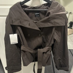 Kort brun trenchcoat från Bershka - Snygg kort brun trenchcoat från Bershka med bred krage, dubbelknäppning och knytskärp i midjan. Jackan har långa ärmar med spänne vid ärmslut och två sidofickor. Storlek M, helt slutsåld🩷