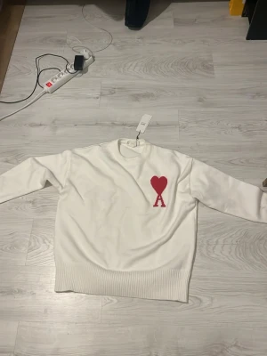 Vit stickad tröja från Ami Paris helt ny - Säljer en vit stickad tröja från Ami Paris med röd hjärtlogga och bokstaven A på bröstet. Tröjan har rund halsringning, långa ärmar och bred ribbstickad mudd vid ärmslut och nederkant. Perfekt för dig som gillar stilrena plagg med en twist.