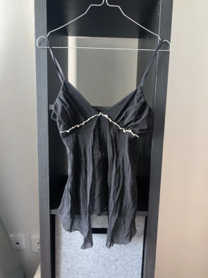 Svart linnetopp med pärldetalj - Säljer en svart linnetopp med tunna axelband och pärldetalj längs bröstet. Toppen har en flowy passform och är tillverkad i ett lätt, skrynkligt material som ger en avslappnad look. Perfekt för dig som gillar stilrena detaljer och enkel design.