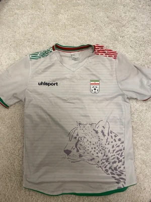 Iran fotbollströja Uhlsport grå - Grå fotbollströja från Uhlsport med Irans landslagsemblem på bröstet och ett stort gepardmotiv på framsidan. Tröjan har korta ärmar, V-ringning och detaljer i rött, grönt och vitt på axlar och krage. 