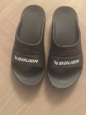 Nya bauer tofflor  - Säljer ett par  gråa tofflor från Bauer med vitt logotryck pga storleken. Modellen är slip-in med öppen tå och tillverkad i mjukt gummi för extra komfort. Perfekta att slänga på sig efter träningen eller till stranden.