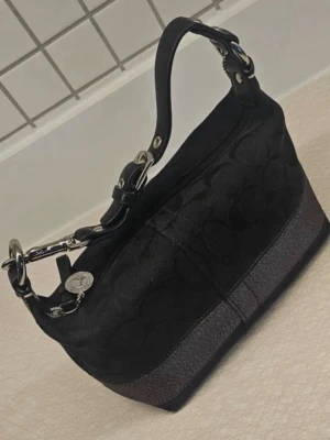 Svart handväska från Coach - Snygg svart handväska från Coach med klassiskt mönster och silverfärgade detaljer. Väskan har en kort axelrem i läder och en dekorativ metallbricka. Materialet är en mix av textil och läder med en mörkgrå botten. Perfekt för dig som gillar stilrena accessoarer.