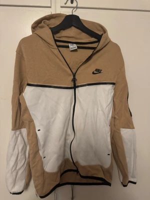 beige och vitt Nike Tech - Snygg nike tech  i beige och vitt med dragkedja framtill och svart Nike-logga på bröstet. Huva och långa ärmar, samt svarta detaljer och fickor framtill. Perfekt för en sportig och avslappnad stil.