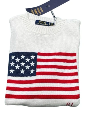 Vit stickad tröja Polo Ralph Lauren - Vit stickad tröja från Polo Ralph Lauren med amerikansk flagga i rött, vitt och blått på framsidan. Rund halsringning och ribbstickade muddar. RL-broderi nertill. Perfekt för dig som gillar klassisk och preppy stil.