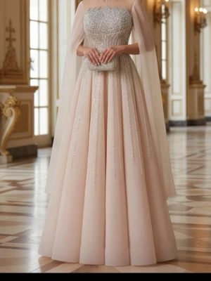 Glittrig långklänning i champagne - Magisk långklänning i champagnefärg med glittriga paljettdetaljer och pärlor upptill. Klänningen har en markerad midja, utsvängd kjol och transparent cape-liknande ärmar. Perfekt för bal eller fest. Materialet är troligtvis polyester och satin. Där jag har markerat med svart jag har lagt till att glittret går neråt