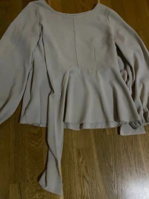Beige blus med knytband - Stilren beige blus med långärmad design och rund halsringning. Blusen har ett dekorativt knytband i midjan och lätt utsvängd nederkant. Tillverkad i ett mjukt, tunt tyg som ger ett snyggt fall.