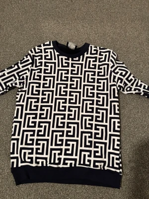 Svartvit mönstrad sweatshirt - En Cool balmain svartvit sweatshirt med grafiskt mönster över hela tröjan. Tröjan har rund halsringning, långa ärmar och ribbade muddar vid ärmslut och nederkant. Perfekt för dig som gillar statement-plagg och streetstyle.