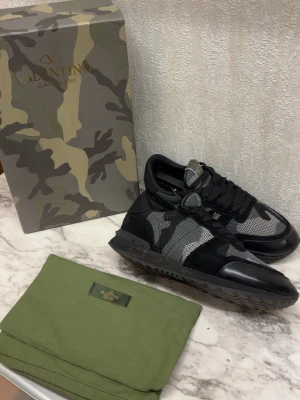 Valentino Garavani camo sneakers 42 - Snygga sneakers från Valentino Garavani med svart och grått camomönster. Skorna har detaljer i mocka och mesh, samt svarta skosnören och sula. Perfekta för dig som gillar streetwear och exklusiv design.