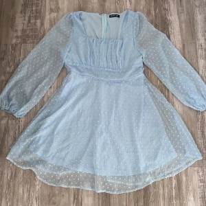Ljusblå kortklänning med prickar - Supersöt ljusblå kortklänning med transparenta långärmar och små prickar. Klänningen har en markerad midja, lätt puffiga ärmar och en flowy kjol. Perfekt för sommarens alla tillfällen! Defekten syns inte när man har på sig klänningen då armen täcker den och hålet är bara på första lagret av tyg☺️