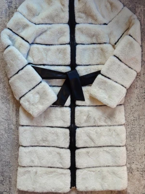 Vit pälsjacka med svarta detaljer - Snygg vit pälsjacka i fuskpäls med svarta horisontella ränder och svart bälte i midjan. Jackan har hög krage och raka ärmar, perfekt för kalla vinterdagar. Ger en lyxig och trendig look.