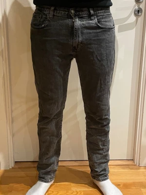 Svarta/gråa slim fit Levi’s jeans - Säljer ett par svarta slim fit jeans med klassisk femficksmodell och knappgylf. Jeansen har en tvättad look och sitter tajt över benen för en modern stil. Perfekta för dig som gillar en enkel men snygg look. Slitet vid ena fickan inget man tänker på! Är enkligen baggy jeans men är försmå för modellen, funkar också så som modellen har! Stolek W32 L32