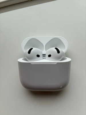 AirPod 4 ANC - Säljer ett par vita trådlösa hörlurar från Apple med stilren design och laddfodral. Hörlurarna har en kompakt form och sitter smidigt i öronen. Perfekta för musik, poddar och samtal. Laddfodralet är också vitt och har en slät yta.