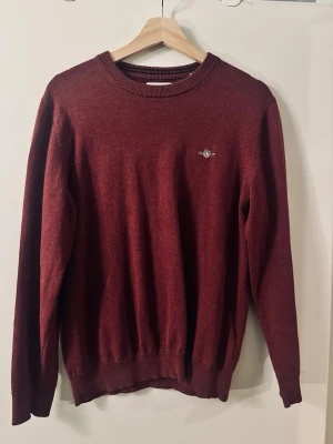 Vinröd stickad tröja från Gant - Vinröd stickad tröja från Gant med rund halsringning och diskret broderad logotyp på bröstet. Tröjan har långa ärmar och ribbade muddar vid ärmslut och nederkant. Perfekt för lager-på-lager och snygg till jeans.