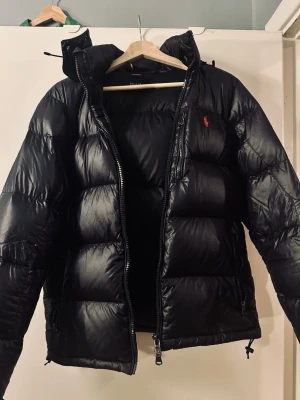 Svart marinblå pufferjacka från Ralph Lauren - Snygg svart marinblå pufferjacka från Ralph Lauren med ikonisk röd logga på bröstet. Jackan har hög krage, dragkedja framtill och justerbar huva. Fylld med vaddering för extra värme och har en klassisk quiltad design. Perfekt för kalla vinterdagar.