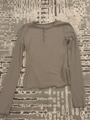Grå ribbad långärmad topp - Snygg grå ribbad topp med långa ärmar och rund hals. Toppen har en knappdetalj framtill som ger en extra touch. Perfekt att styla med jeans eller kjol för en chill look.