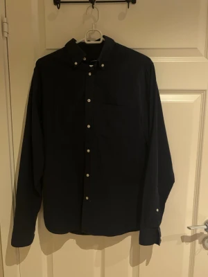 J.lindberg skjorta  - Stilren mörkblå skjorta från j.lindberg med klassisk krage och vita knappar framtill. Skjortan har lång ärm och en bröstficka, perfekt för en clean och enkel look. Perfekt för sommaren runt hörnet