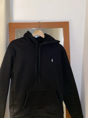 Polo hoodie - Svart hoodie från Polo Ralph Lauren med klassisk huva och dragsko. Framtill finns en stor magficka och den ikoniska broderade loggan i vitt på bröstet. Perfekt för en chill och stilren look. Materialet är mjukt och bekvämt.