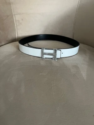 Vitt skärp med silverfärgat H-spänne - Stilrent vitt bälte med ett stort H-format spänne i silverfärg. Bältet har en slät yta och insidan är svart. Perfekt accessoar för att lyfta din outfit med en modern touch.