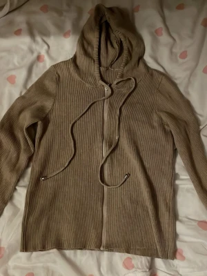Beige ribbad hoodie med dragkedja - Säljer en beige ribbad hoodie med dragkedja framtill och justerbar huva med snören. Hoodien har långa ärmar och är tillverkad i ett mjukt, stretchigt tyg som ger en avslappnad look. Perfekt för en chill och stilren outfit.