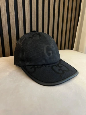 Svart keps från GG med GG-mönster - Snygg svart keps från Gucci med diskret GG-mönster över hela kepsen. Kepsen har böjd skärm och justerbart spänne bak i läder. Tillverkad i ett slitstarkt tygmaterial som ger en exklusiv känsla. Perfekt accessoar för en trendig streetstyle.