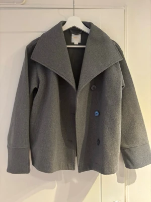 Grå ullrock med bred krage - Stilren grå rock med bred krage och två knappar framtill. Rak passform och långa ärmar med markerade muddar. Tillverkad i ett ullblandat material som ger en klassisk och tidlös look.