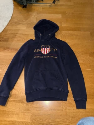 Mörkblå hoodie från GANT - Snygg mörkblå hoodie från GANT med broderad logga och sköld på bröstet. Klassisk känguruficka framtill, dragsko i huvan och ribbade muddar. Perfekt för en avslappnad och sportig stil. Materialet känns mjukt och bekvämt.