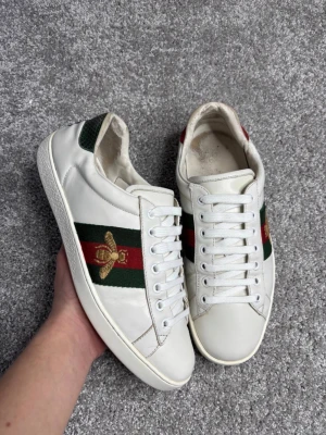 Gucci ace bee - Säljer ett par vita Gucci sneakers i läder med klassiska gröna och röda ränder på sidorna samt en broderad guldfärgad bi. Skorna har vita snören och grön detalj vid hälen. Perfekta för dig som vill ha en lyxig och trendig look.
