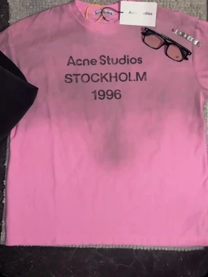 Rosa Acne Studios t-shirt - Säljer en rosa t-shirt från Acne Studios med tryck 'Acne Studios STOCKHOLM 1996' på bröstet. Klassisk rund hals och korta ärmar. Perfekt för dig som gillar streetwear och vill ha något från ett ikoniskt svenskt märke.