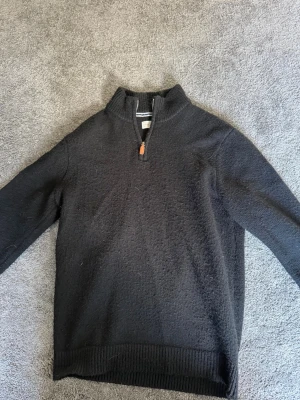 Svart halvzip-tröja från Pier One - Svart stickad tröja från Pier One med halv dragkedja och ribbad krage. Tröjan har långa ärmar och ribbade muddar vid ärmslut och nederkant. Perfekt för lager-på-lager och enkel att matcha med jeans eller chinos.