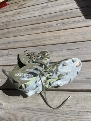 Adidas Predator fotbollsskor grön/svart - Säljer ett par Adidas Predator fotbollsskor i grön och svart med coolt geometriskt mönster och silvriga detaljer. Skorna har snörning och vit sula med dobbar för bra grepp på planen. Perfekta för dig som vill sticka ut på matchen!
