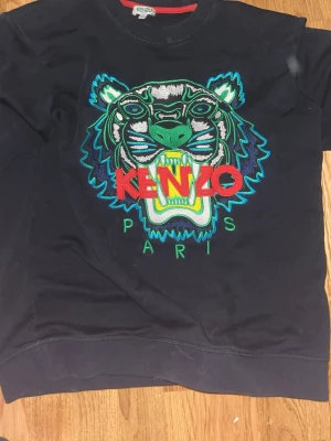 Svart Kenzo hoodie med tigertryck - Svart hoodie från Kenzo med ikoniskt broderat tigerhuvud i grönt, blått och vitt samt röd Kenzo-logga och texten Paris. Tröjan har rund halsringning och ribbade muddar. Materialet är mjukt och bekvämt, perfekt för en cool streetstyle-look.