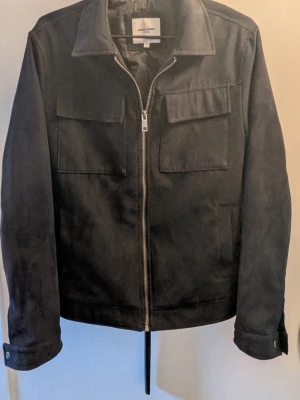 Svart overshirt från Jack & Jones - Stilren svart overshirt från Jack & Jones med två stora bröstfickor och dragkedja framtill. Klassisk krage och raka ärmar med knappdetalj vid mudden. Materialet ser ut att vara mjukt och matt, perfekt för lager på lager.