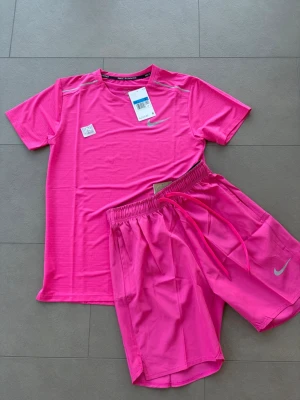 Nike set  - Säljer ett fräscht rosa träningsset från Nike med Dri-Fit teknologi. Setet består av en t-shirt och shorts, båda i lätt och snabbtorkande material. Perfekt för gymmet eller löprundan. Båda plaggen har Nike-logga och elastisk midja på shortsen.