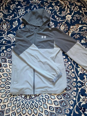 Grå vindjacka från Under Armour - Snygg tvåfärgad grå vindjacka från Under Armour med huva och dragkedja framtill. Jackan har logga på bröstet och textdetalj på ärmen. Perfekt för blåsiga dagar och sportiga outfits.