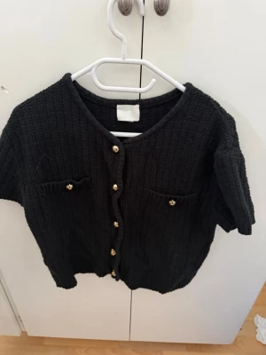 Svart kofta med guldknappar H&M - Stilren svart kofta från H&M med kort ärm och snygga guldknappar framtill. Koftan har två små fickor på bröstet och är stickad i ett mjukt material. Perfekt för att lyfta din outfit med en klassisk touch.