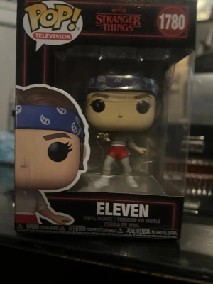 Funko Pop Eleven Stranger Things - Säljer en Funko Pop vinylfigur av Eleven från Stranger Things, nummer 1780. Figuren har blå bandana, röda shorts och vita skor. Perfekt för samlare eller fans av serien. Levereras i originalförpackning med cool design.