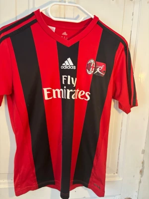 AC Milan matchtröja Adidas röd/svart (2013-14) - Säljer en klassisk AC Milan matchtröja från Adidas i rött och svart med vita detaljer. Tröjan har klubbmärke, sponsorlogga och tre svarta ränder på axlarna. Tillverkad i lätt och ventilerande material, perfekt för fotbollsträning eller match.