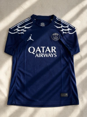 PSG matchtröja blå Jordan - Säljer en blå Paris Saint-Germain matchtröja från Jordan med vita detaljer och vingmönster på axlarna. Tröjan har PSG-logga, Jordan-logga och Qatar Airways-tryck på bröstet. Perfekt för fotbollsträning eller att bära som supporter.