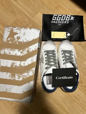 Golden Goose vita sneakers  - Golden Goose sneakers i vitt läder med grå mockadetaljer, grå stjärna på sidan och blå häl. Grå skosnören och vintage-inspirerad sula. Klassisk låg modell med GGDB/SSTAR-tryck på sidan. Levereras med originalkartong och certifikat.
