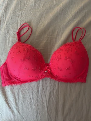 Rosa push-up bh - Snygg rosa push-up bh med broderade blommor, spetsdetaljer och en söt rosett med liten pärla framtill. Dubbel axelband och vadderade kupor ger extra stöd och form. Perfekt för dig som vill ha något både bekvämt och fint under kläderna. Bh-n är i en skrik rosa färg, perfekt till sommaren!