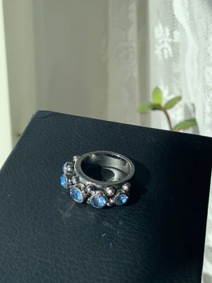 Silverring med blå stenar - Unik silverfärgad ring med flera runda, ljusblå stenar runt framsidan av bandet. Ringen har en chunky design med små silverkulor mellan stenarna för extra detalj. Perfekt för dig som gillar statement-smycken och vill sticka ut.
