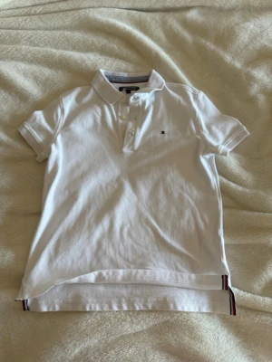 Vit pikétröja från Tommy Hilfiger - Klassisk vit pikétröja från Tommy Hilfiger med korta ärmar, krage och knappar framtill. Liten broderad logga på bröstet och diskreta ränder i nederkant. Perfekt till jeans eller shorts för en clean look.