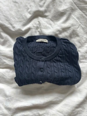 Mörkblå kabelstickad kofta - Snygg mörkblå kabelstickad kofta från Basic Cph med rund halsringning och knappar framtill. Långärmad modell med ribbade muddar och nederkant. Perfekt att slänga över en t-shirt eller klänning för en extra touch.